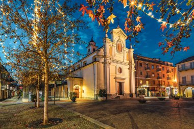 Küçük kasaba meydanındaki San Giovanni Battista Kilisesi. Akşamları Alba, Piedmont, Kuzey İtalya 'da Noel aydınlatmalı ağaçlarla çevrili..