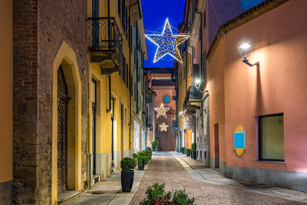 View of narrow cobblestone evening street among old buildings with Christmas illumination in Alba, Piedmont, Northern Italy.