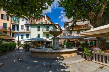 DOLCEACQUA, İtalya - 27 Temmuz 2021: Dolceacqua 'da küçük meydandaki ağaçların altındaki çeşme ve açık hava restoranları - Fransa sınırındaki kasaba, ünlü ve popüler turizm beldesi.
