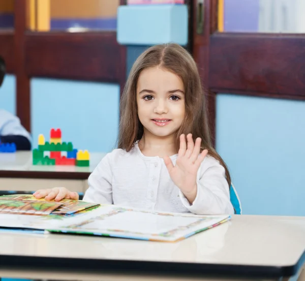 Mädchen mit Popup-Buch winkt am Schreibtisch im Kindergarten — Stockfoto