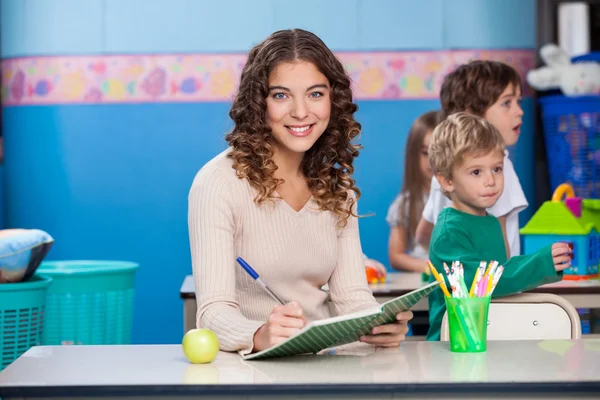 Teacher Writing In Book With Children Playing In Background — Stock Photo, Image