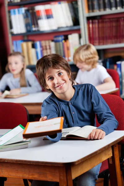 Happy Schoolboy dando livro enquanto sentado na mesa na biblioteca — Fotografia de Stock