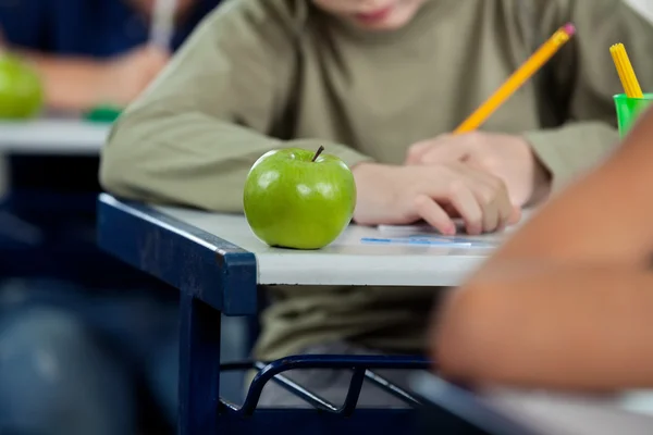 Schooljongen schrijven in boek met apple aan balie — Stockfoto