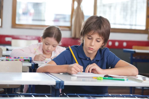 Menino escrevendo notas com colega de classe no fundo — Fotografia de Stock