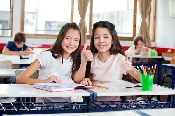 Glückliche Schulmädchen gestikulieren mit den Daumen nach oben am Schreibtisch — Stockfoto