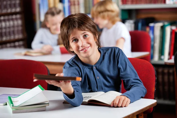 Söt skolpojke ger bok medan du sitter vid bordet i biblioteket — Stockfoto