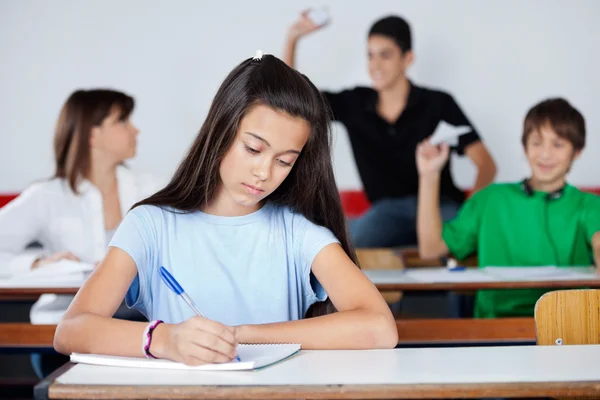 Schoolgirl Writing At Desk While Classmates Playing — Stock Photo, Image