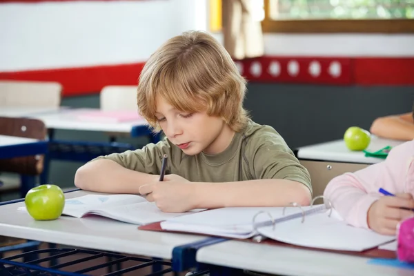 Schoolboy Writing On Book In Classroom Stock Photo