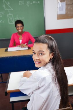 arka planda gülümseyen öğretmen ile masaya oturan kız öğrenci
