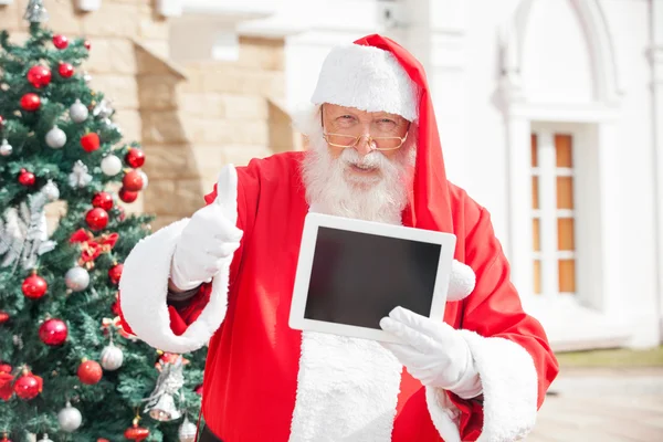 Άγιος Βασίλης χειρονομώ thumbsup κρατώντας ψηφιακή δισκίο — Φωτογραφία Αρχείου