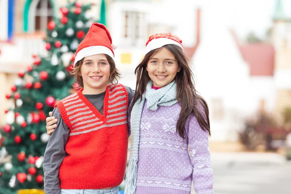 Vrienden in santa hat staande tegen kerstboom — Stockfoto