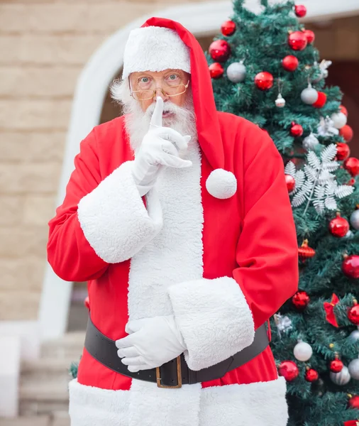 Santa Claus con el dedo en los labios contra el árbol de Navidad — Foto de Stock