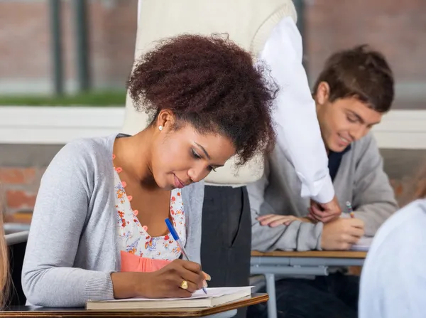 Student schriftelijk examen terwijl leraar uitleggen klasgenoot in backg — Stockfoto