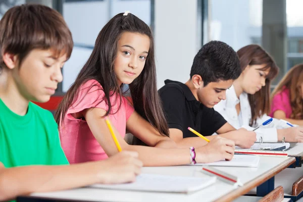 Teenage Girl Sitting With Classmates Writing At Desk Stock Photo