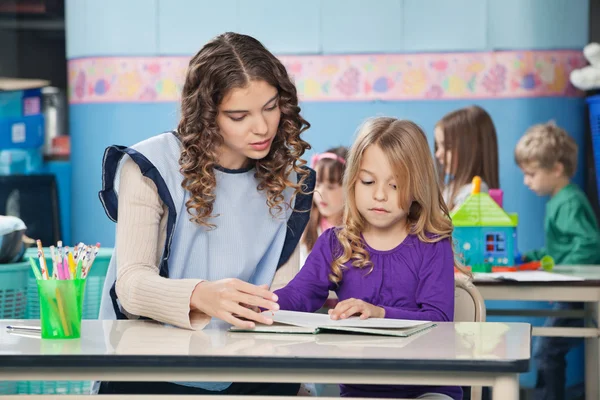Lehrerin und Mädchen lesen Buch mit Kindern im Hintergrund — Stockfoto