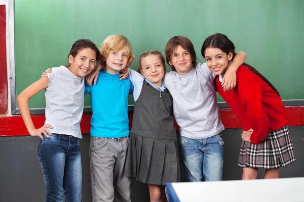 Gelukkig schoolkinderen met armen rond permanent samen in de klas — Stockfoto