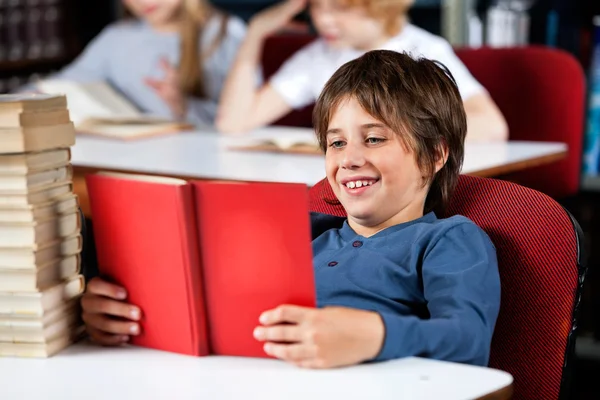 Skolpojke leende medan du läser boken på bordet i biblioteket — Stockfoto