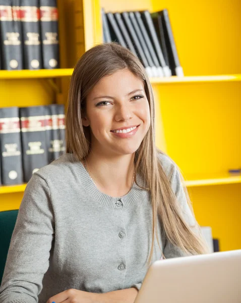 Student med laptop leende mot bokhyllan i college library — Stockfoto