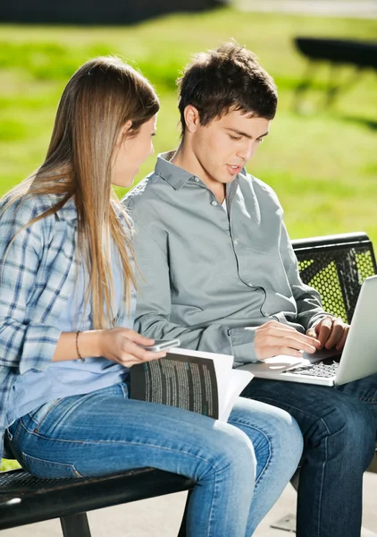 Amici con computer portatile e libro seduto in Campus — Foto Stock