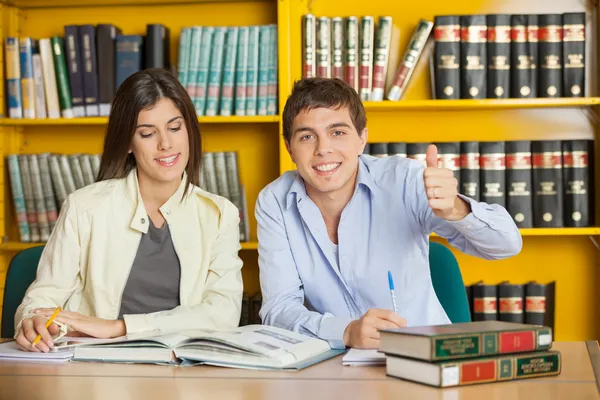 Estudante Gesturing Thumbsup enquanto amigo leitura livro na biblioteca — Fotografia de Stock