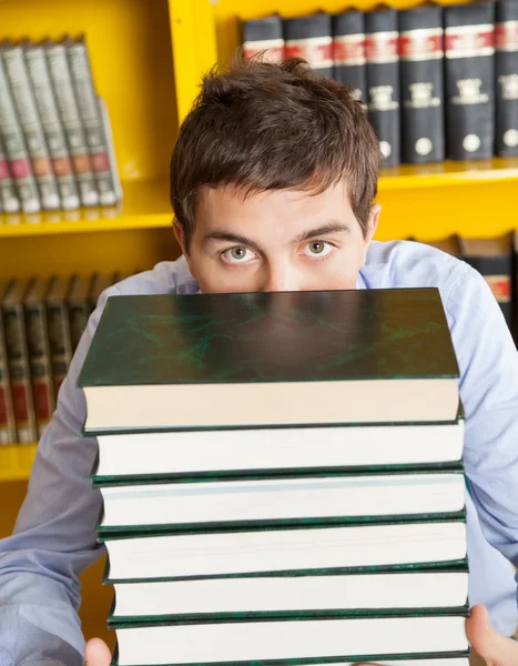 Mannelijke student gluren over gestapelde boeken in bibliotheek — Stockfoto