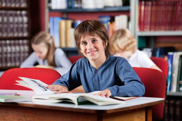 Skolpojke leende med böcker på bord i biblioteket — Stockfoto