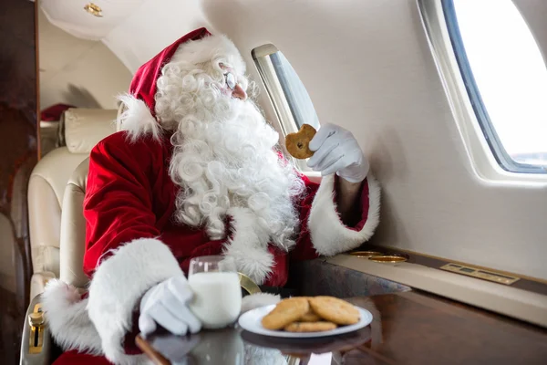 Weihnachtsmann hält Plätzchen, während er durch das Fenster des Privatjets blickt — Stockfoto