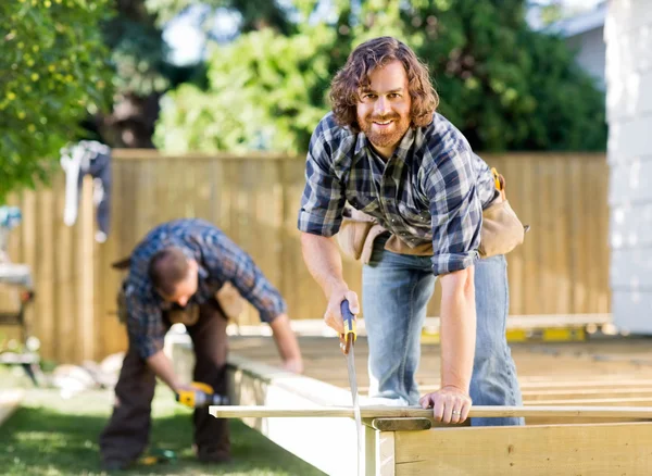 Carpenter Cutting Wood With Saw While Coworker Drilling At Site — Stock Photo, Image