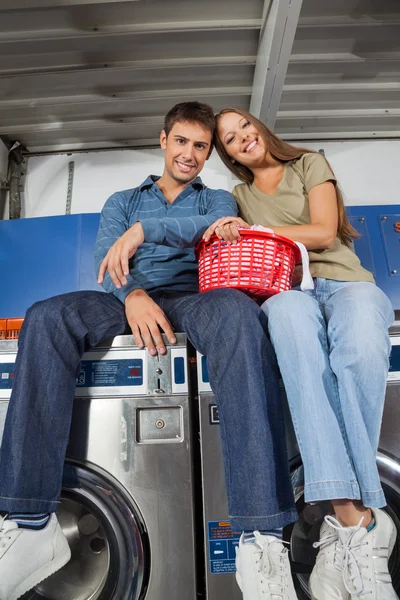 Пара с корзиной для белья сидя на стиральных машинах — стоковое фото