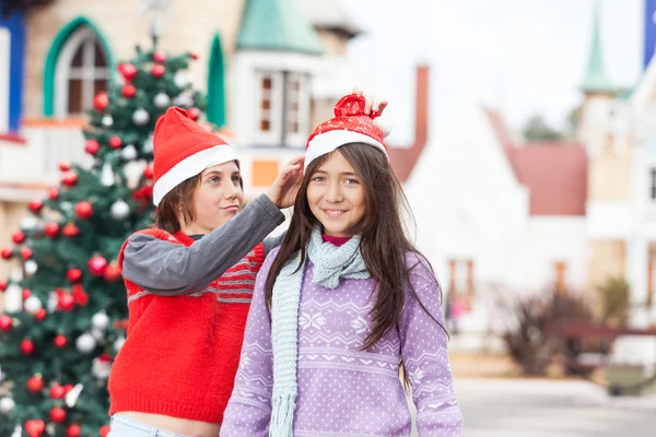 Chica sonriendo mientras amigo poner santa sombrero en su cabeza —  Fotos de Stock