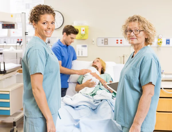 Enfermeiras sorridentes em pé contra casal com bebê no fundo — Fotografia de Stock
