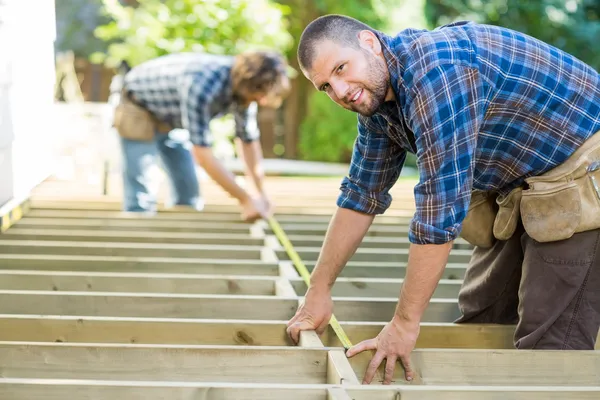 Měření dřeva s páskou, zatímco spolupracovníka mu asistovala Carpenter — Stock fotografie