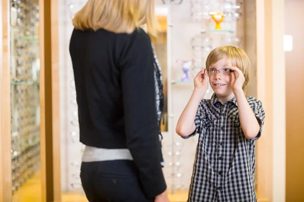 Мальчик смотрит на мать, примеряя очки в магазине — стоковое фото