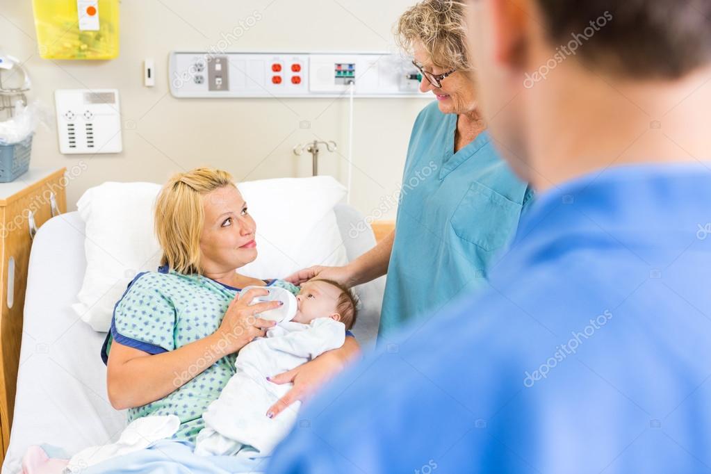 Nurse Looking At Woman Feeding Milk To Babygirl In Hospital