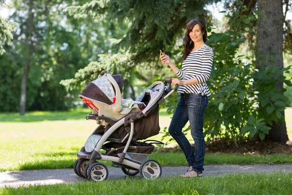 Madre con cellulare e carrozza a piedi nel parco — Foto Stock