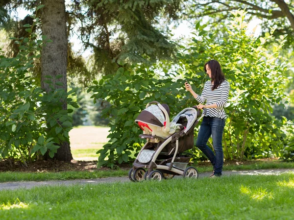 Mujer con el transporte del bebé usando el teléfono celular en el parque —  Fotos de Stock