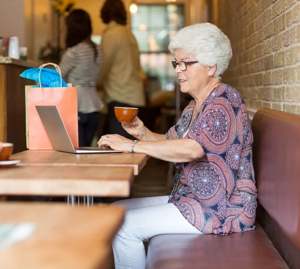 कॅफेरीमध्ये कॉफी घेत असताना लॅपटॉप वापरणे वरिष्ठ महिला — स्टॉक फोटो, इमेज