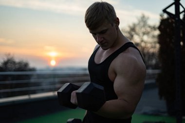 Beyaz kaslı beyaz adam günbatımının arka planında dambılları kaldırıyor. İrade, motivasyon ve sağlıklı yaşam tarzı kavramı