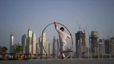 Cyr Wheel artist wearing white Arabic clothing cloth on Dubai cityscape background during sunset
