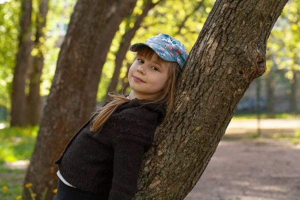 一个可爱的7岁小女孩头戴棒球帽站在树边的户外肖像 — 图库照片