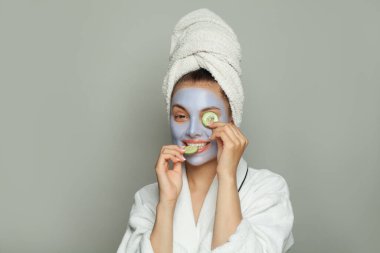 Yeşil salatalık tutan nemlendirici maskeli mutlu spa kadını. Mavi kozmetik maskeli kadın model. Yüz bakımı ve cilt bakımı kavramı