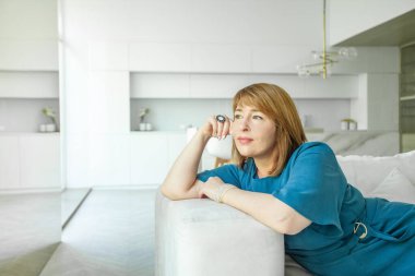 Orta yaşlı bir kadın modern oturma odasındaki koltukta dinleniyor, dinleniyor ve stres yok, başka tarafa bakıyor, düşünüyor.