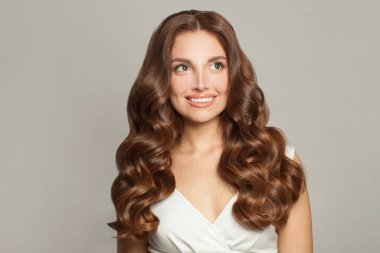Güzel kahverengi saçlı, makyajlı, sağlıklı kıvırcık saçlı, gri arkaplanına bakan güzel bir kadın.