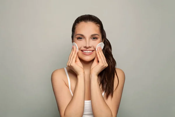 Healthy fresh woman removing makeup from her face with cotton pads and smiling. Beauty model cleaning her face with cotton swab pad on grey background. Skin care and beauty concept.