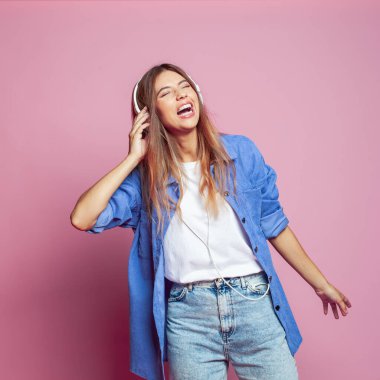 Mutlu genç bir kadın, harika bir şarkıyla rahatlamış, geniş bir gülüşü var, kulaklıklara dokunuyor, müzik dinliyor, mavi bir gömlek giyiyor, pembe stüdyo duvarında izole edilmiş, dans etmek istiyor..