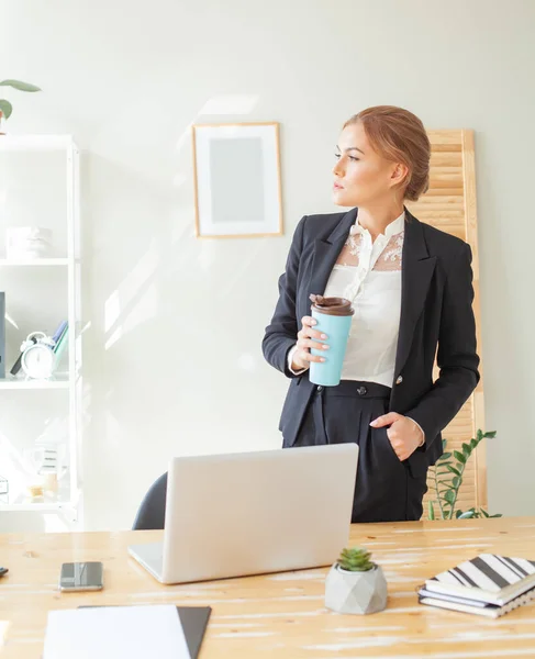 头脑清醒的年轻女性穿着时髦休闲装 站在办公室的工作岗位上 一边举着咖啡杯 一边微笑着看着远方 — 图库照片