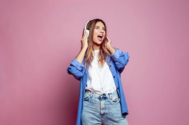 Kulaklıklı, müzik dinleyen ve pembe arka planda şarkı söyleyen güzel genç bir kadın.
