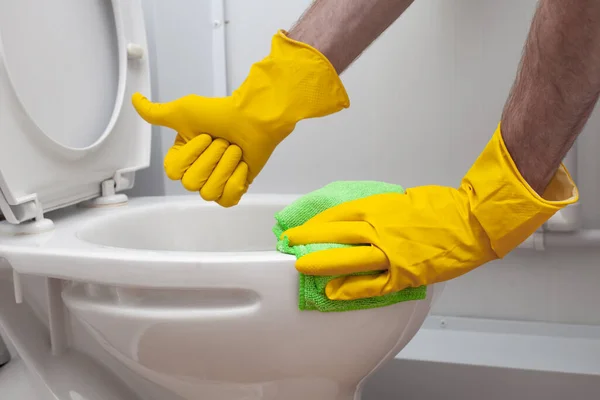 Male hand in yellow rubber glove showing thumb up and cleaning white toilet bowl
