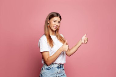 Pembe arka planda baş parmak gösteren neşeli genç bir kadının portresi.