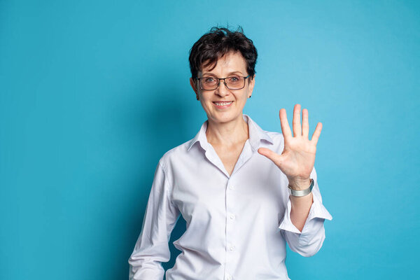 Portrait of smiling middle adged woman in glasses showing five fingers against blue studio wall background